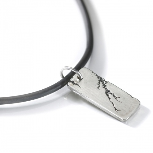 A distinctive chunky silver men’s pendant hangs off a black rubber cord. The silver dog tag is solid matt silver with contrasting black crack.